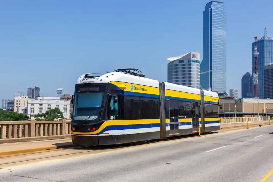White and yellow DART streetcar on bridge in Dallas, Texas with skyscrapers in background.